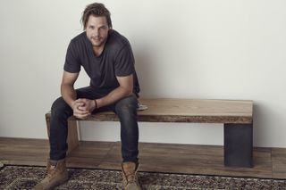 Mats Christéen wearing a dark t-shirt, jeans and brown boots sitting on a black and wooden bench in a room with light grey walls, wood flooring and a dark coloured patterned rug