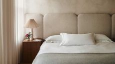 Close-up of a neutral bed with an extended headboard that matches the pinkish-beige color of the walls. There is white bedding with three throw cushions. 