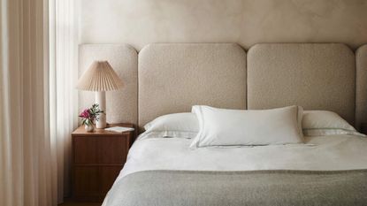 Close-up of a neutral bed with an extended headboard that matches the pinkish-beige color of the walls. There is white bedding with three throw cushions. 