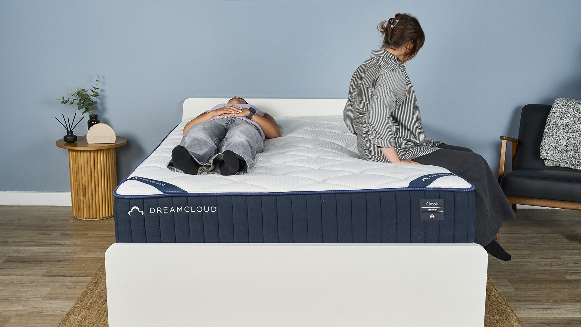 One Tom&#039;s Guide tester lies on her back on the DreamCloud Classic inside a blue-painted bedroom while another gets into bed, lies on her side and then lies on her other side