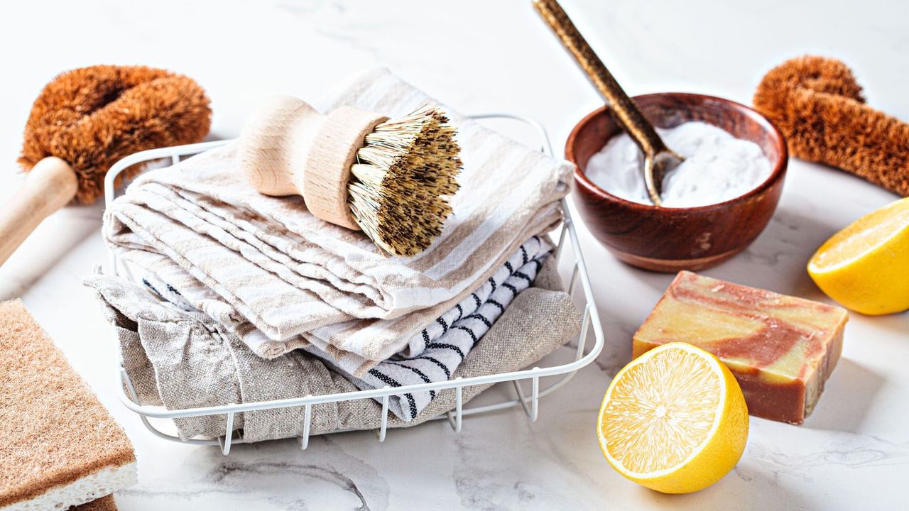 A pile of cleaning cloths with a scrubbing brush beside a owl of baking soda and a cut open lemon