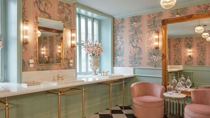 Pastel green and peach bathroom with triple sinks and seating with Botanize Heritage Plaster Pink Wallpaper, which has vertical columns of pale green foliage on pink background 