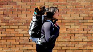 A woman in front of a brick wall carries a backpack with a dog in it