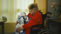Elderly woman sat in wheelchair cradles robot like a baby. 