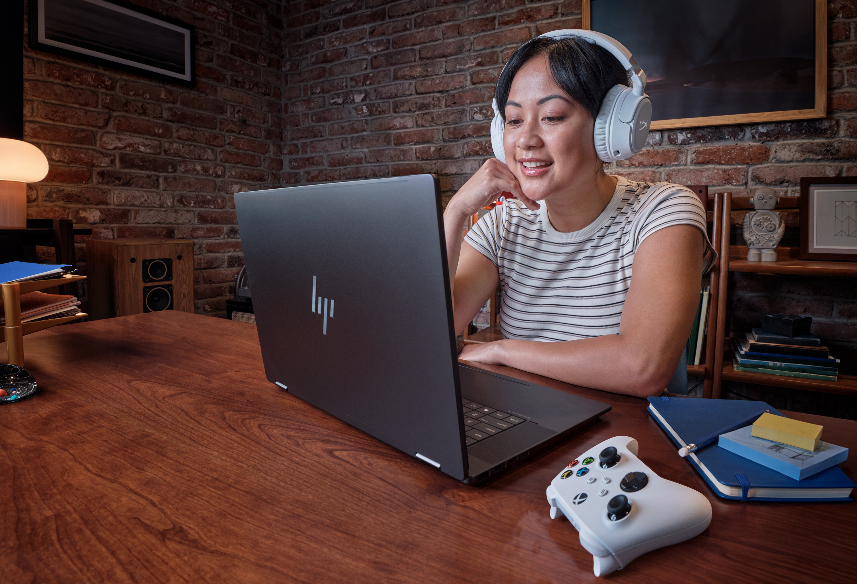 A woman sits at a wood table wearing headphones with an Xbox controller beside the HP OmniBook X Flip 14".