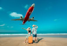 A plane flies low over two tourists, waving from a beach and holding luggage.