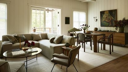 Image of an open concept living room and dining area with white wood-paneled walls, a wooden dining table, and an L-shaped sofa.