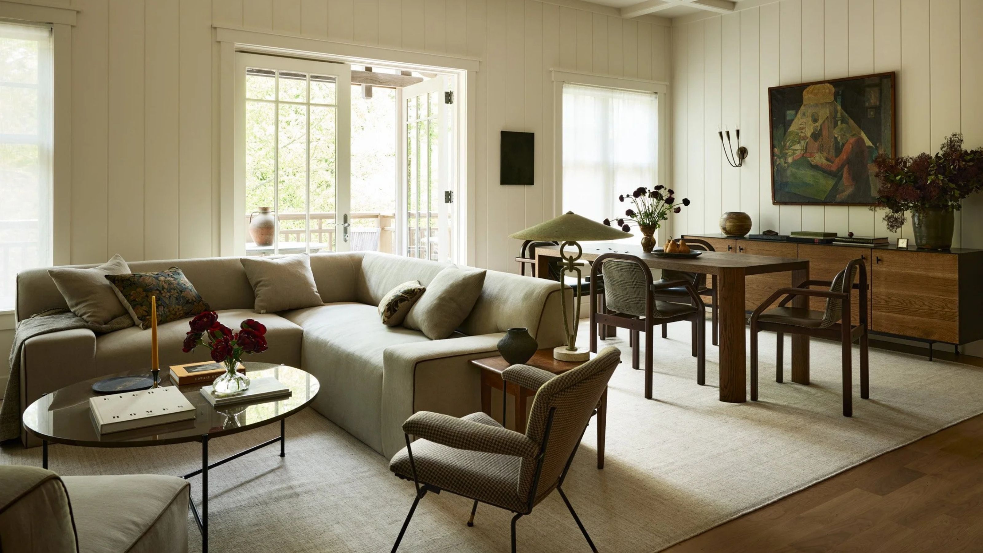 Image of an open concept living room and dining area with white wood-paneled walls, a wooden dining table, and an L-shaped sofa. 