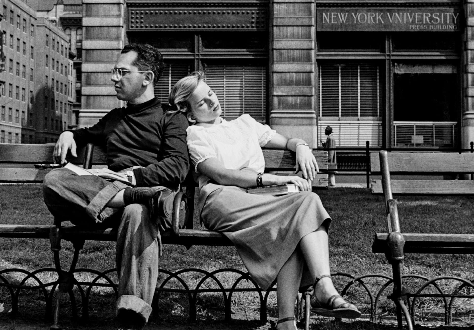 A woman rests her head on a man's shoulder on a Washington Square Park bench, eyes closed, while he gazes away into the middle distance with a book in his lap.