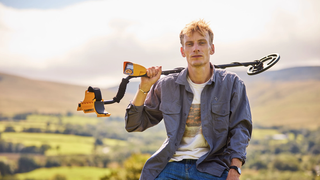 Charlie Cooper leans on a fence in the countryside with a metal detector resting on his shoulder in &quot;Charlie Cooper&#039;s Myth Country&quot;