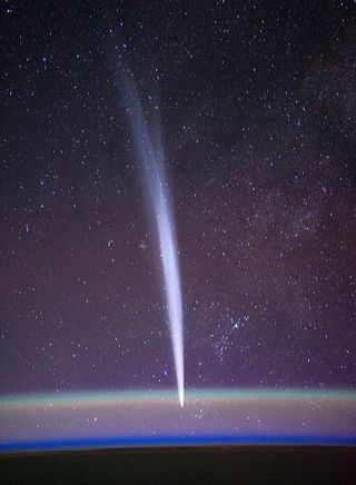 the white streak of a comet stretches downard as it dips below the curved layer of Earth's atmosphere, bulging from bleow, as the starry sky of space shines above.