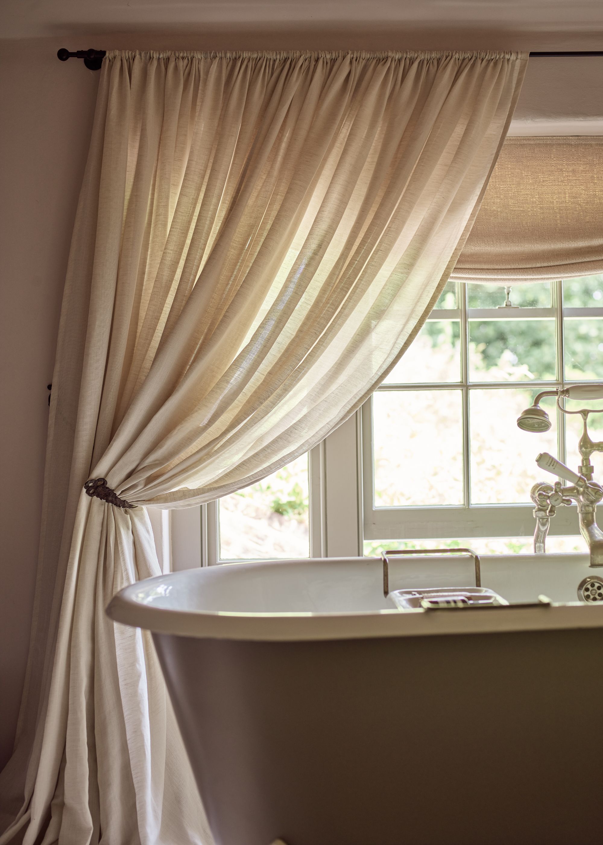 Corner of a bathroom with a roll-top bath and rod pocket sheer curtains in front of a traditional window.
