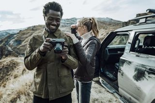 Two people outside in the hills with cameras and a car