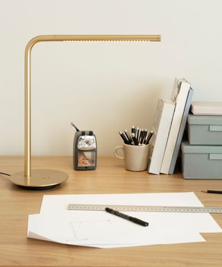 A modern, slim right angled gold table lamp on a wooden desk with paper, a pencil sharpener, and blue stacked shallow storage boxes on it
