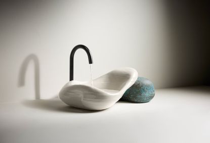 Daniel Arsham Kohler sink made of 3D printed white ceramic resting on an oxidised brass stone-shaped object, with water coming out of a minimalist black tap