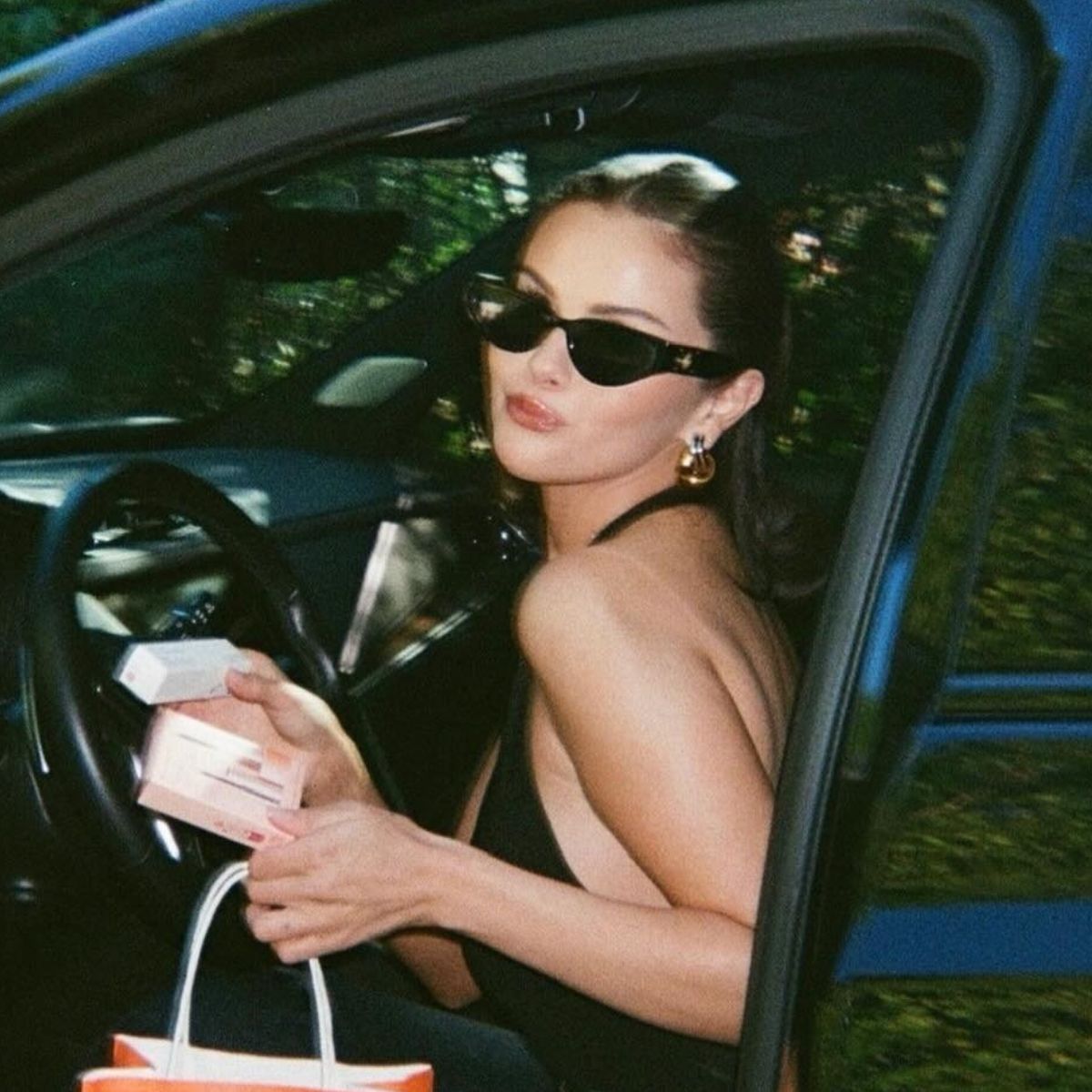 An image of Selena Gomez holding two Rare Beauty products and an Ulta shopping bag while posing in the open door of a car.