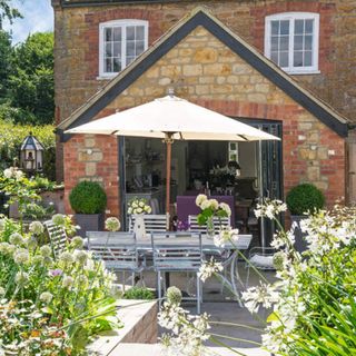 Cottage exterior wall with a large open door into the garden, patio seating area with table and chairs and plants in large wooden raised beds. Pub Orig. A former sloping garden, redesigned and terraced cottage garden in a village in Dorset, home of Judith and Michael Rust.