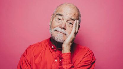 Portrait of senior man in red shirt with head in hand against pink background