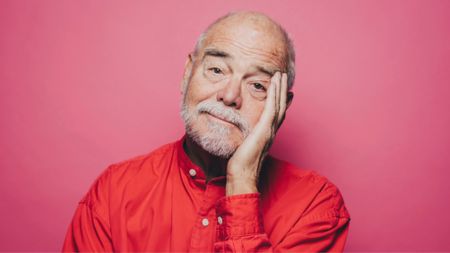 Portrait of senior man in red shirt with head in hand against pink background
