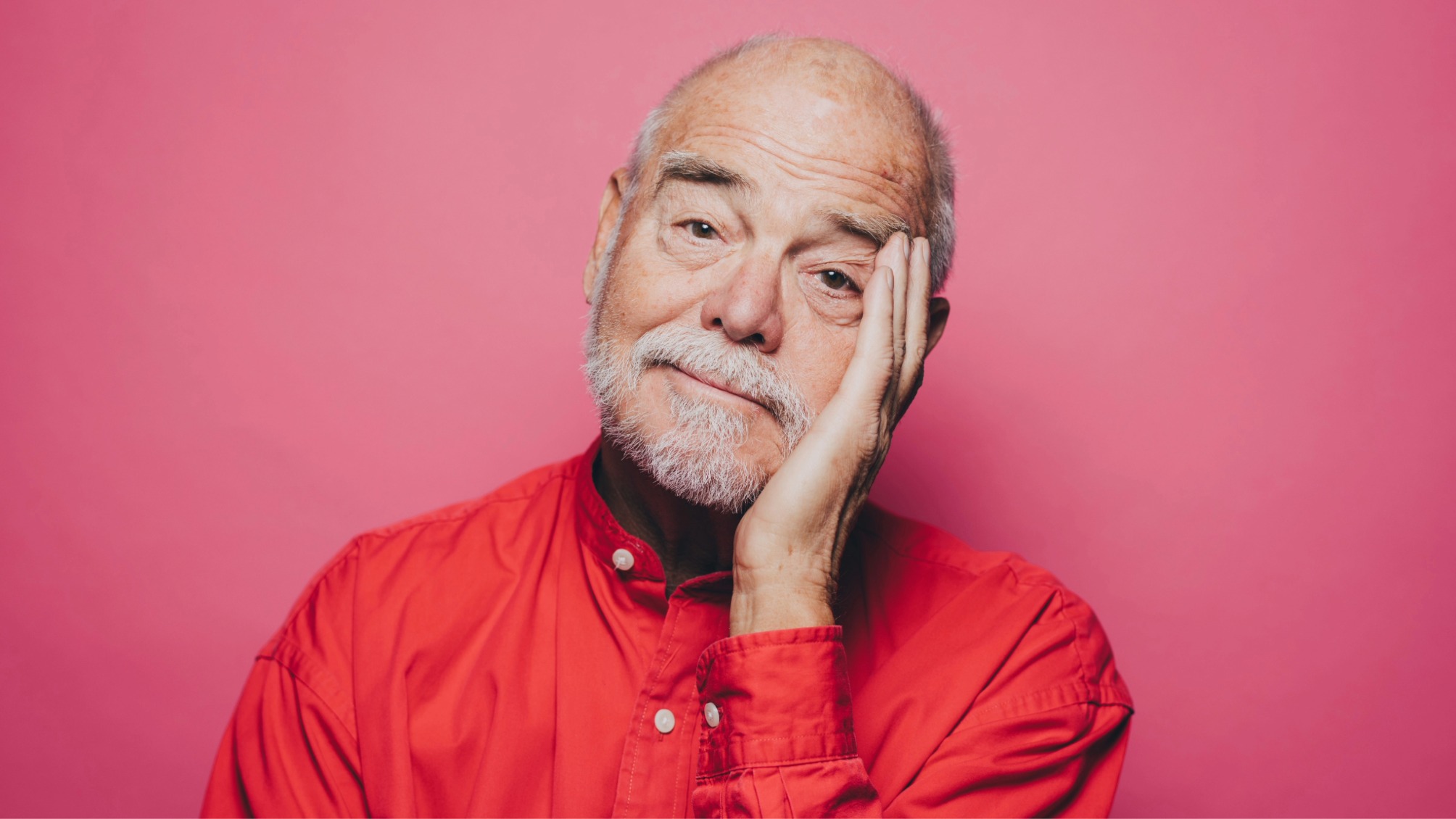 Portrait of senior man in red shirt with head in hand against pink background