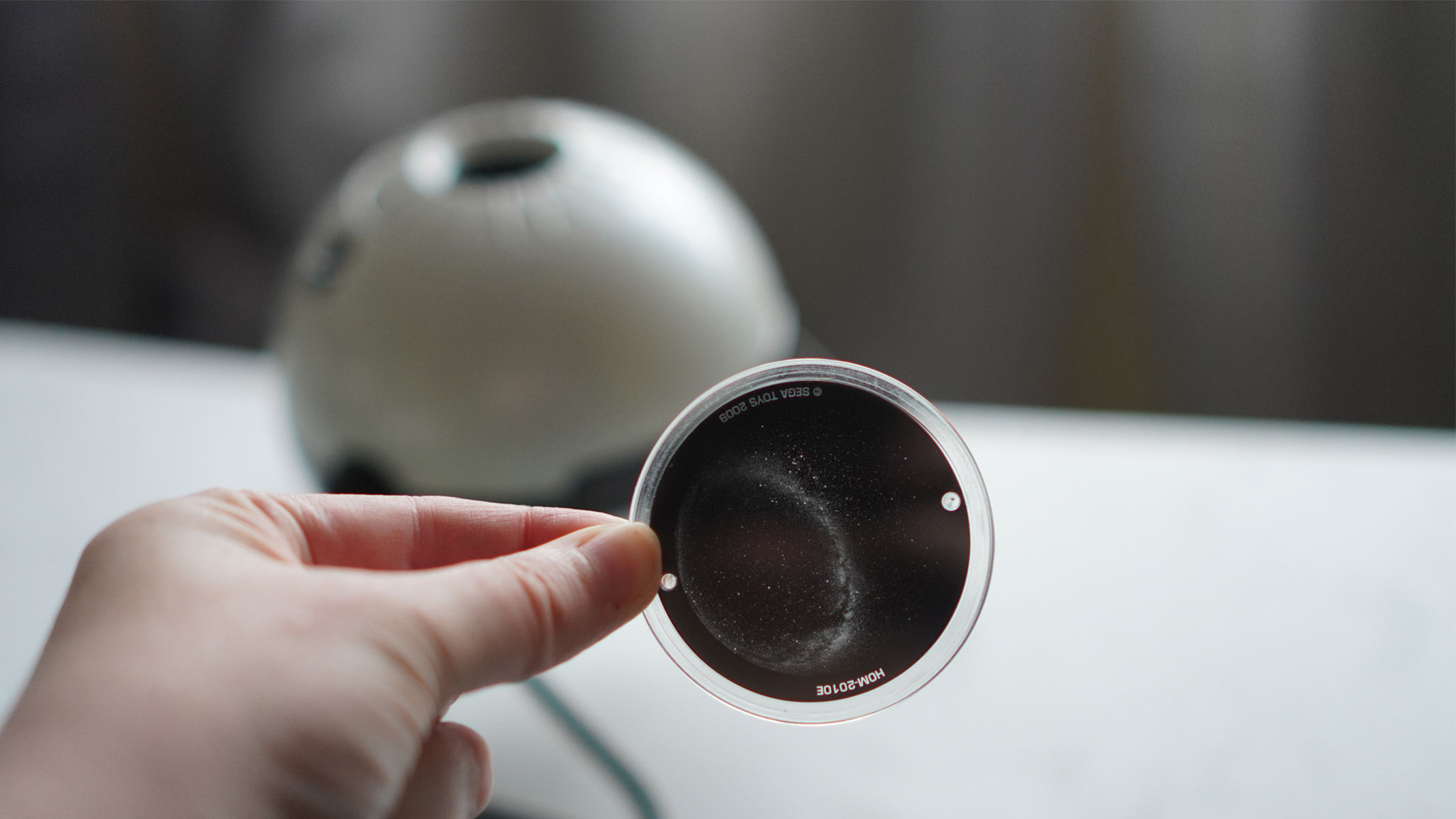 A hand is holding a dark disk in front of a white sphere on a white surface.