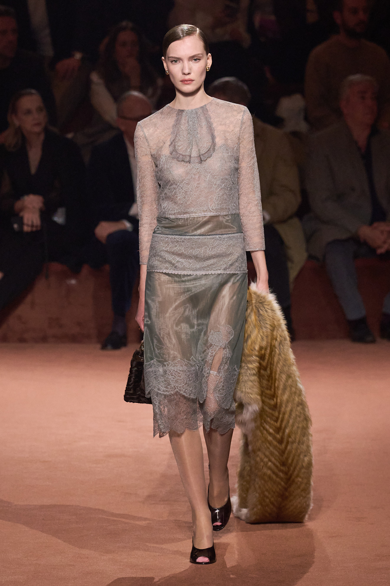 Fur accessory trend shown in Fendi&#039;s fall winter 2025 collection in a photo of a model walking down the runway wearing a sheer gray lace top with a matching slip skirt, dark brown peep toe pumps, a furry Fendi bag, and a fur scarf that&#039;s dragging on the ground