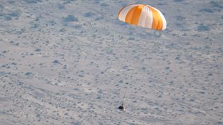 a small capsule falls toward earth under an orange and white parachute.