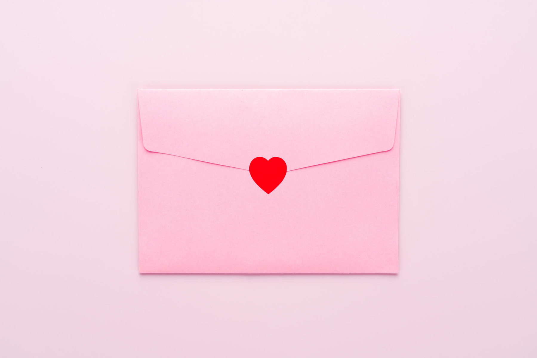 A white envelope is sealed with a red heart. Against a pink background.
