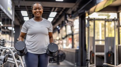 A woman holds a pair of dumbbells at her sides in the gym. She is smiling and we see the blurred outline of machinery behind her.