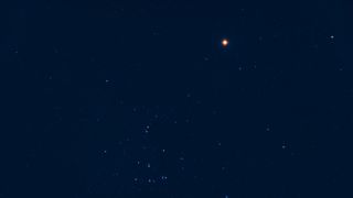 An image of the night sky showing Mars as a red star-light object close ot a number of blue-white stars against a dark blue sky.