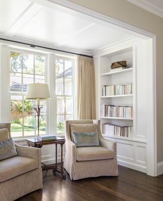 Two neutral armchairs in window with side table and reading lamp and bookshelves in white panelling