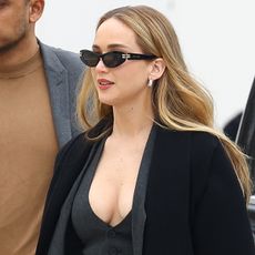 Jennifer Lawrence wearing a gray tailored waistcoat, blazer, and skinny pants with pumps and a black coat at the Dior show in Paris.