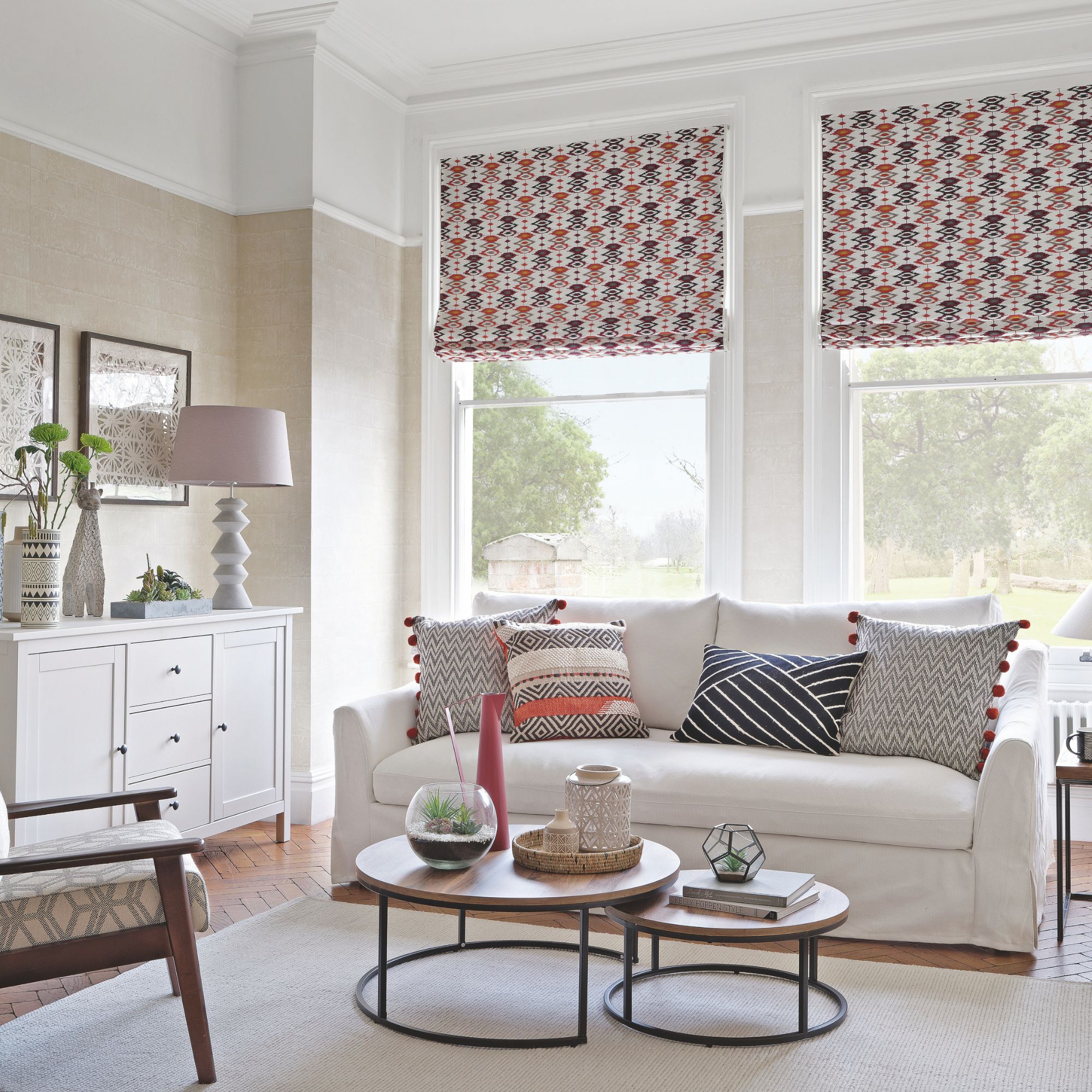 Neutral living room, with light sofa and patterned cushions, coffee table nest, and patterned blinds on the two large windows