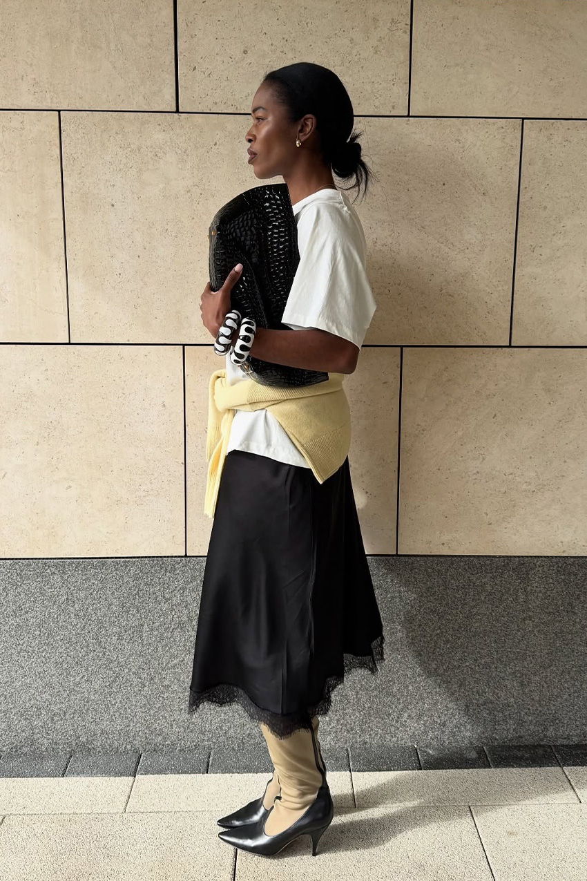 How to wear a slip skirt is shown in a photo of a woman&#039;s slip skirt outfit where she&#039;s standing outside in front of a concrete wall wearing a white oversized t-shirt with a butter-yellow sweater tied around the waist styled with a black lace slip skirt, black and tan two-tone knee-high boots, white and black bangles, and a black east-to-west clutch bag