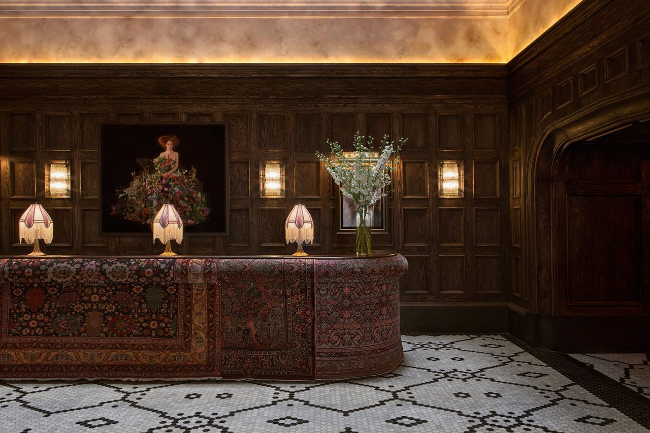 An hotel lounge features warm light, Art Deco table lamps, rugs, wooden wall paneling, and tiled flooring.