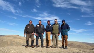 five men stand in a barren arctic landscape under a blue sky dotted with wispy clouds