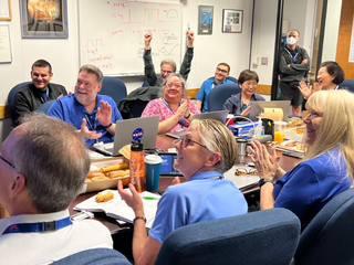 A bunch of people, many of whom are wearing blue, cheer in a conference room.
