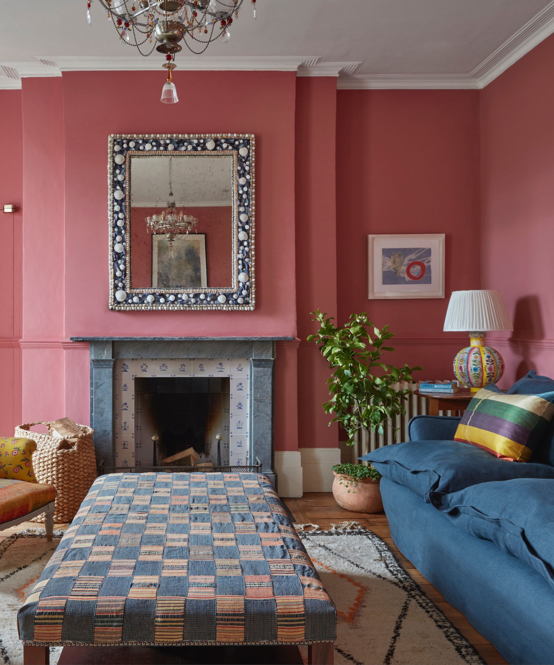 pastel living room ideas, mid-pink living room with denim couch, patchwork style coffee table, shell mirror, chandelier, rug, bright lamp, artwork, rattan basket, plant