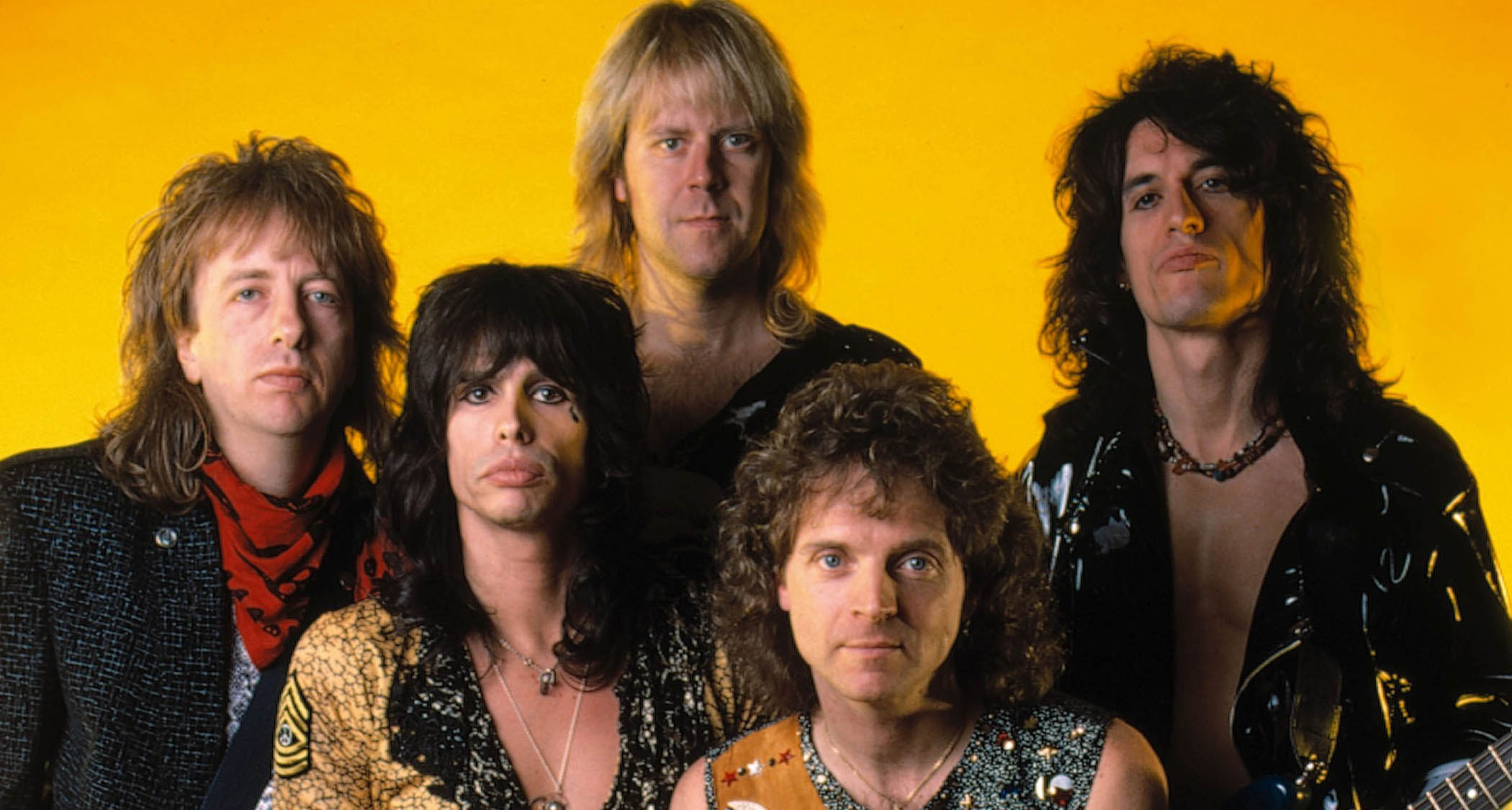 Aerosmith circa 1985/'86, photographed against a yellow backdrop: (L-R) Tom Hamilton, Steven Tyler, Brad Whitford, drummer Joey Kramer, and Joe Perry