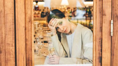 A pensive young woman alone with a glass of champagne looks out of a restaurant window