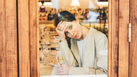 A pensive young woman alone with a glass of champagne looks out of a restaurant window
