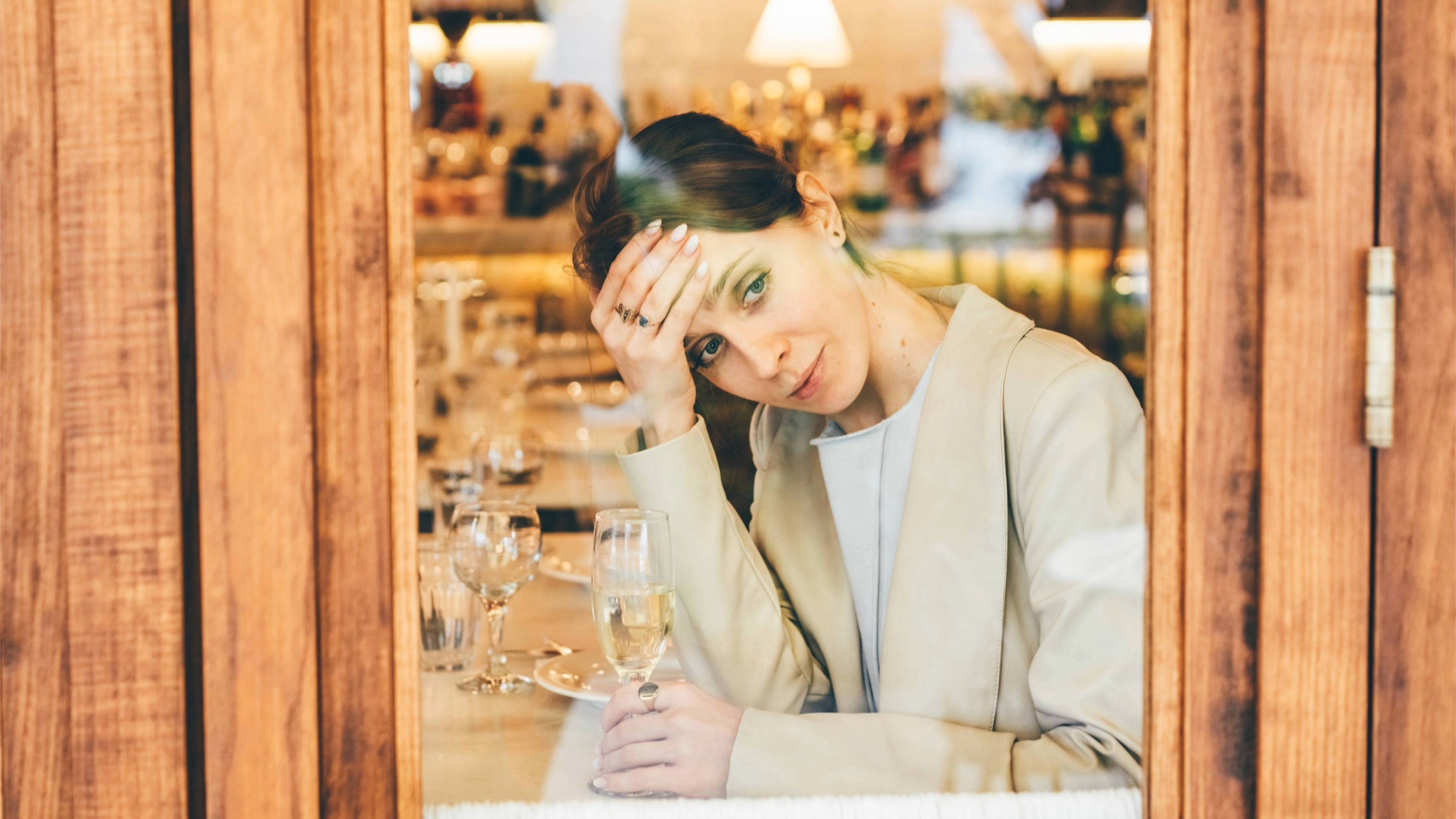 A pensive young woman alone with a glass of champagne looks out of a restaurant window