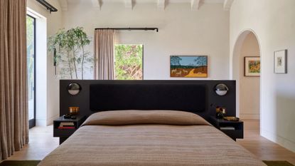 A bright white bedroom with an arched doorway, a colorful framed painting, beige curtains on black rods, a potted tree, a bed with a black frame in the middle of the room, patterned cream sheets and attached side tables