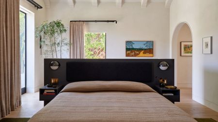 A bright white bedroom with an arched doorway, a colorful framed painting, beige curtains on black rods, a potted tree, a bed with a black frame in the middle of the room, patterned cream sheets and attached side tables