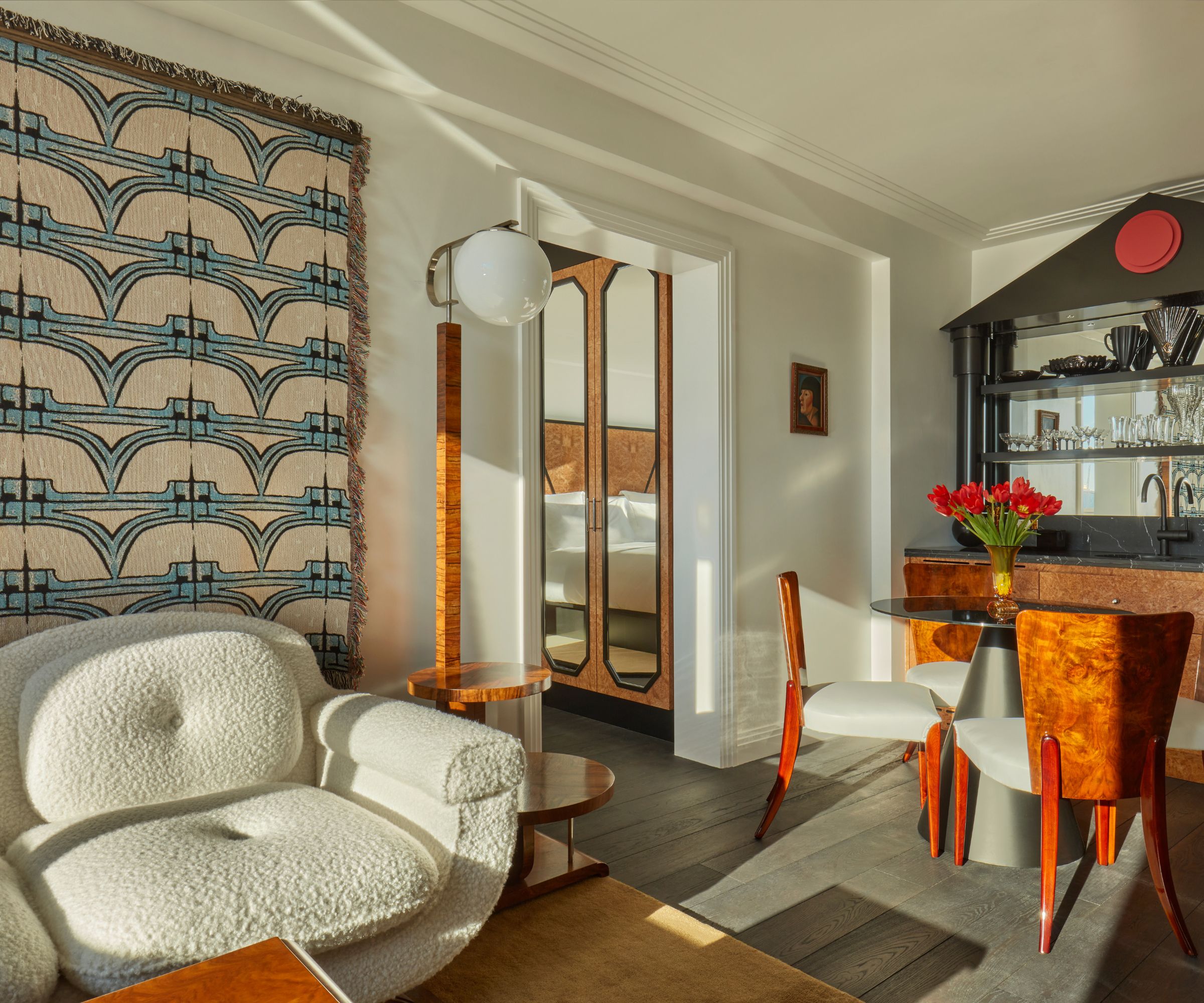 lounge area of a suite by the Lore Group with a boucle couch, wall hanging tapestry, a large bar area with a table and chairs with contrasting metal and burl wood finishes