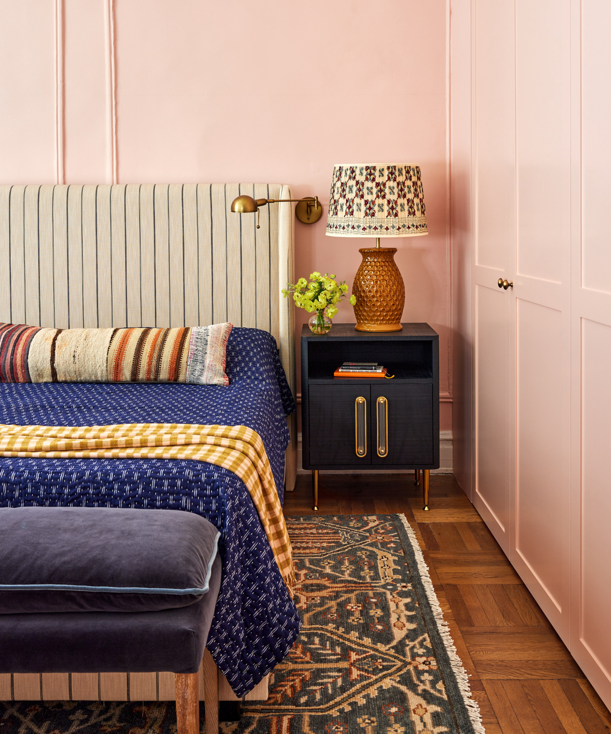 colors that go with light pink, bedroom with light pink walls, stripe upholstered bed, indigo blue blanket, navy nightstand, navy ottoman, vintage rug, hardwood floor, wall light, patterned shade on table lamp, stripe bed pillow, turmeric checked blanket, wardrobes