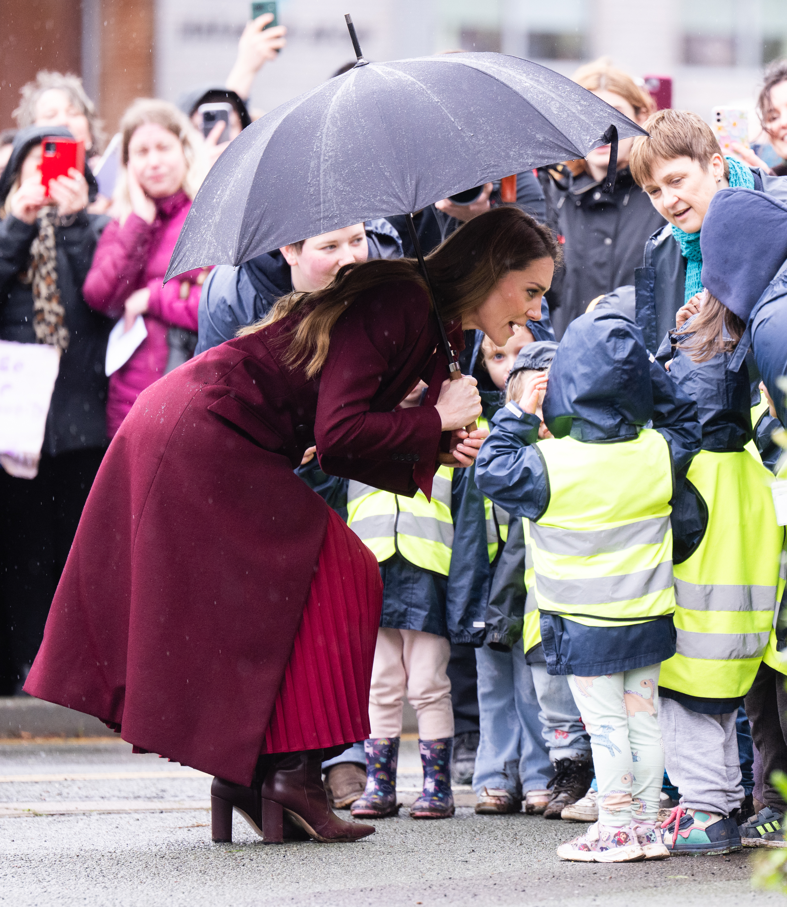 Princess Kate wearing a red coat, holding an umbrella and leaning down to talk to kids