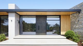 large modern porch with metal roof stretching across entrance and rest of property, timber clad below with inset spotlights and small wall lights on dividing wall and external wall