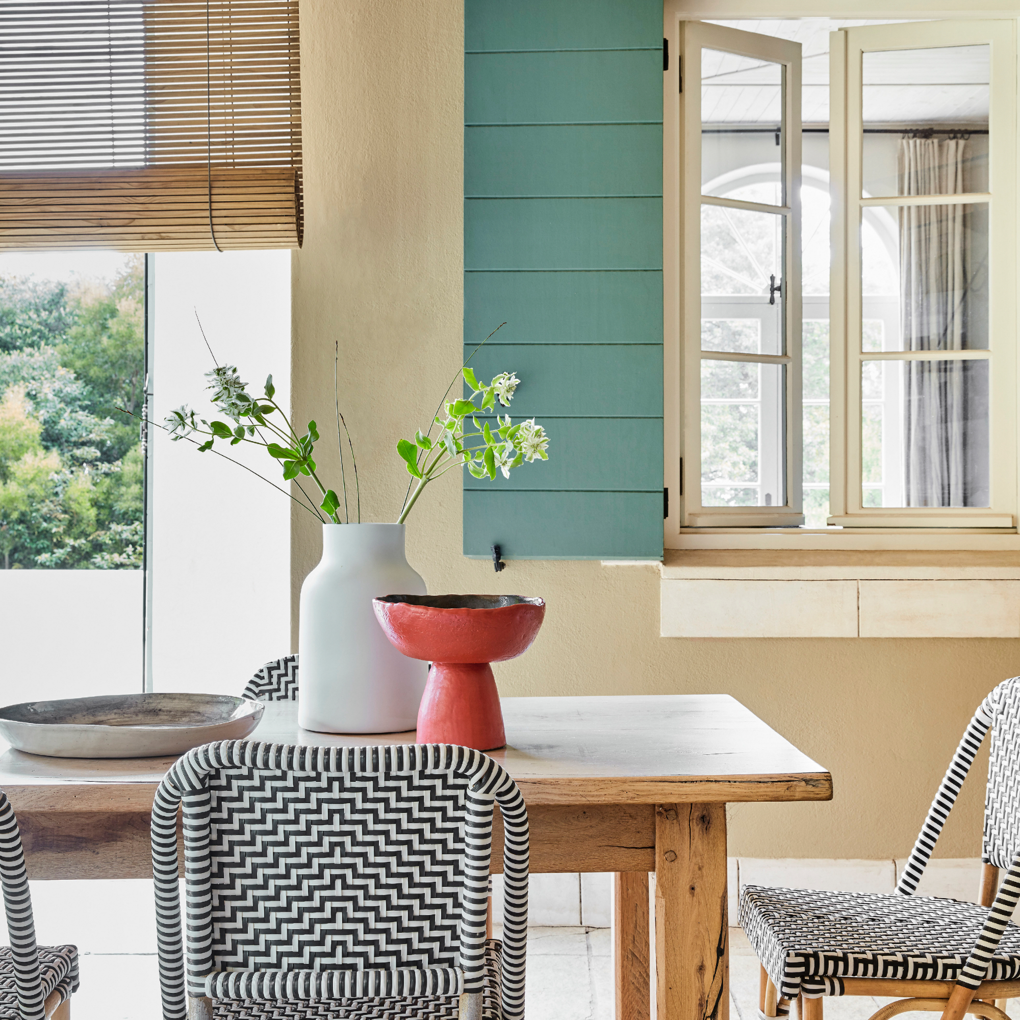 dining room with view outside with roller blind for privacy, painted shutters, black and white rattan chairs, red vase on table, white vase