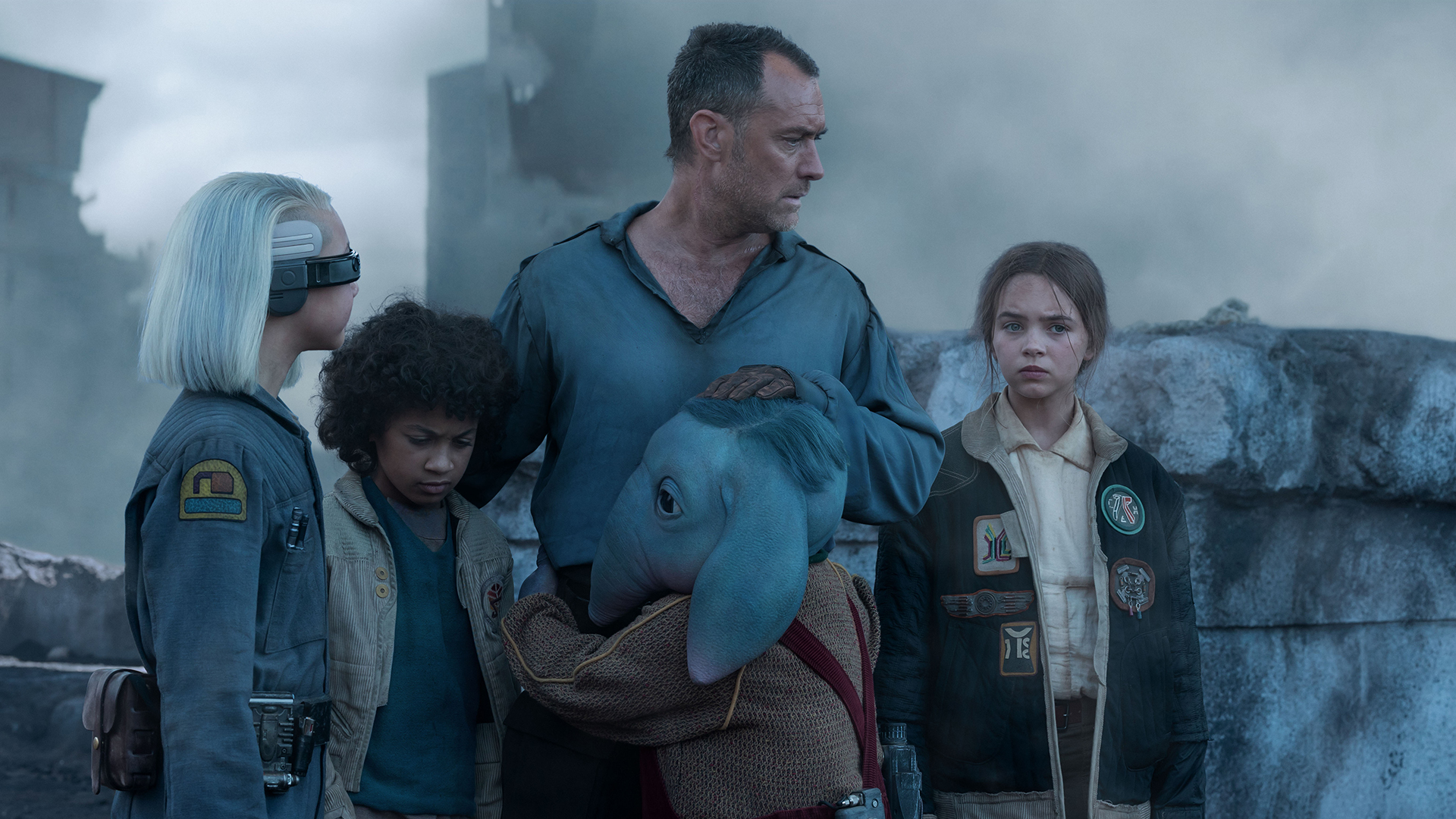 Still from Star Wars: Skeleton Crew TV series. The human man Jod Na Nawood is standing in the middle, surrounded by four children. They look a bit shocked and worn out. From left to right: KB (girl with an asymmetrical white bob and wearing a visor across her eyes), Wim (human boy with dark curly hair) is sadly looking downwards, Neel ((blue elephant-like alien boy) is hugging Jod's waist, and Fern (human girl).